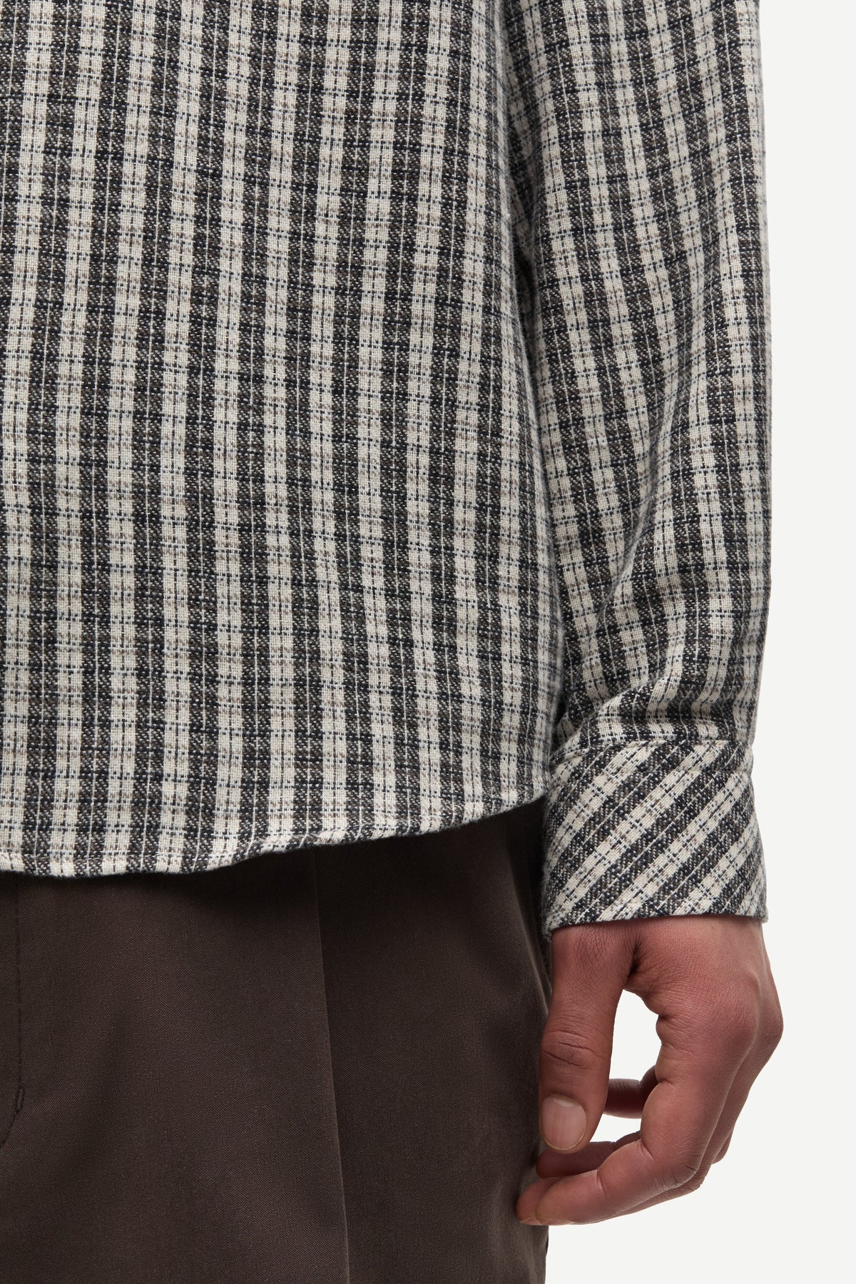 Close-up of a person wearing a long-sleeve plaid shirt in black, white, and gray, paired with dark brown trousers, against a plain light background.