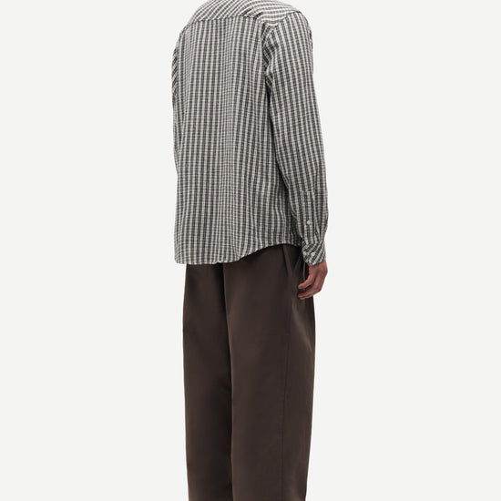 A man shown from the back wearing a black and white vertically striped button-down shirt and dark brown trousers, standing against a plain light gray background.