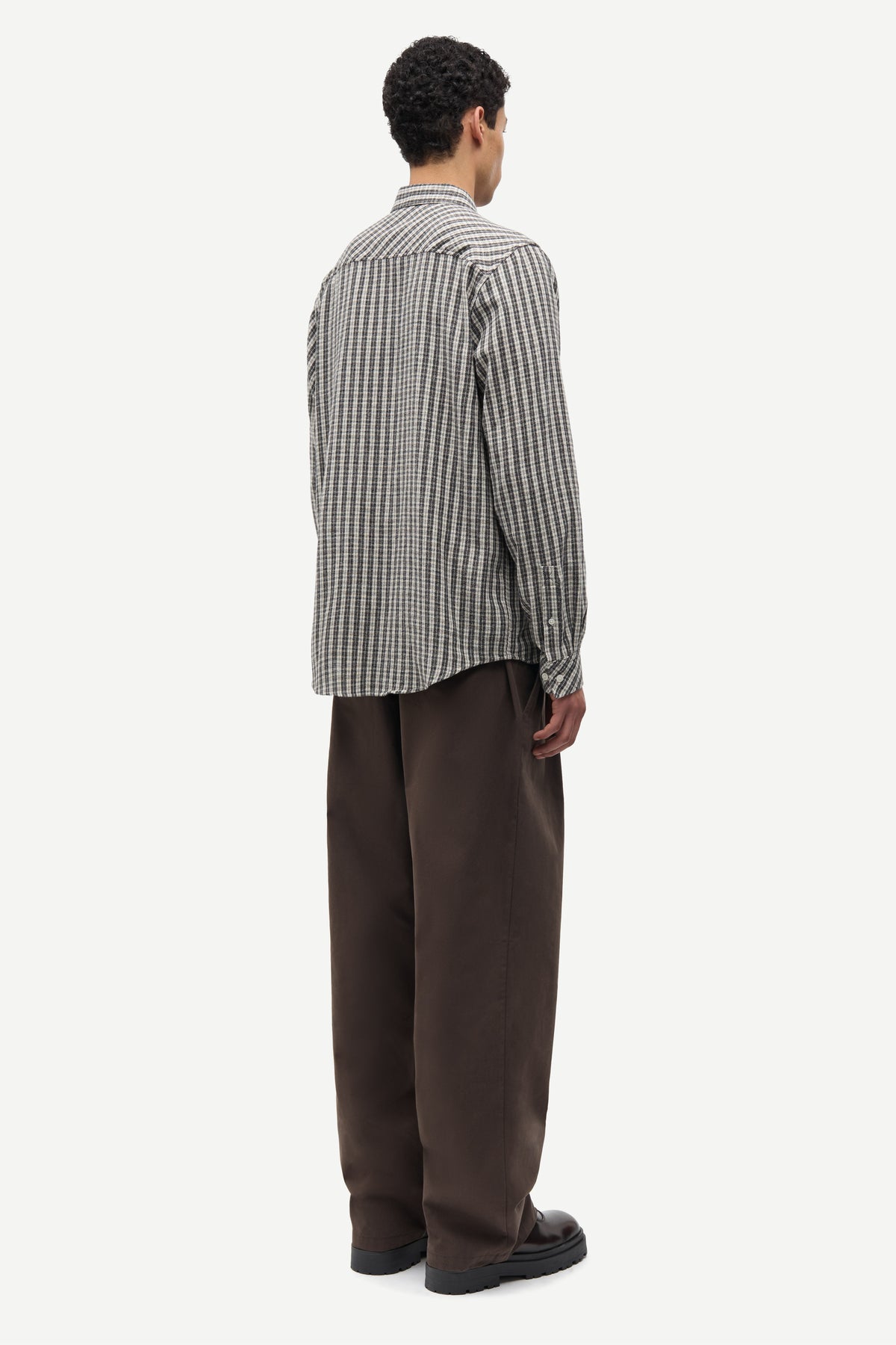 A man shown from the back wearing a black and white vertically striped button-down shirt and dark brown trousers, standing against a plain light gray background.