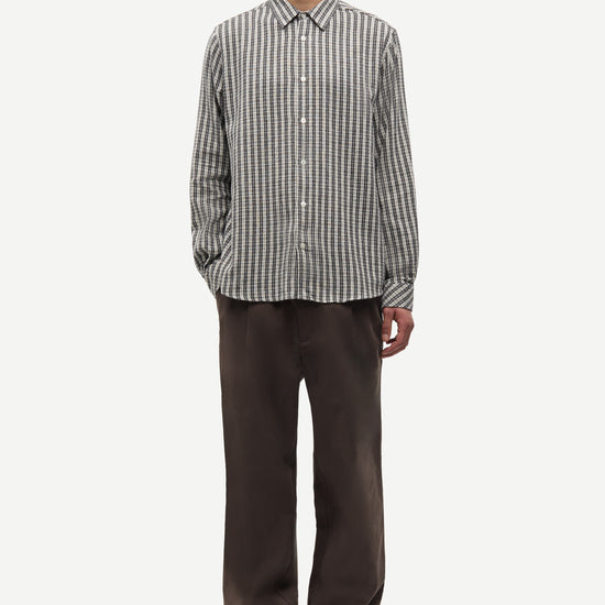 A man wearing a long-sleeve button-up shirt with vertical gray and white stripes, paired with dark brown trousers and dark brown shoes, standing against a light gray background.