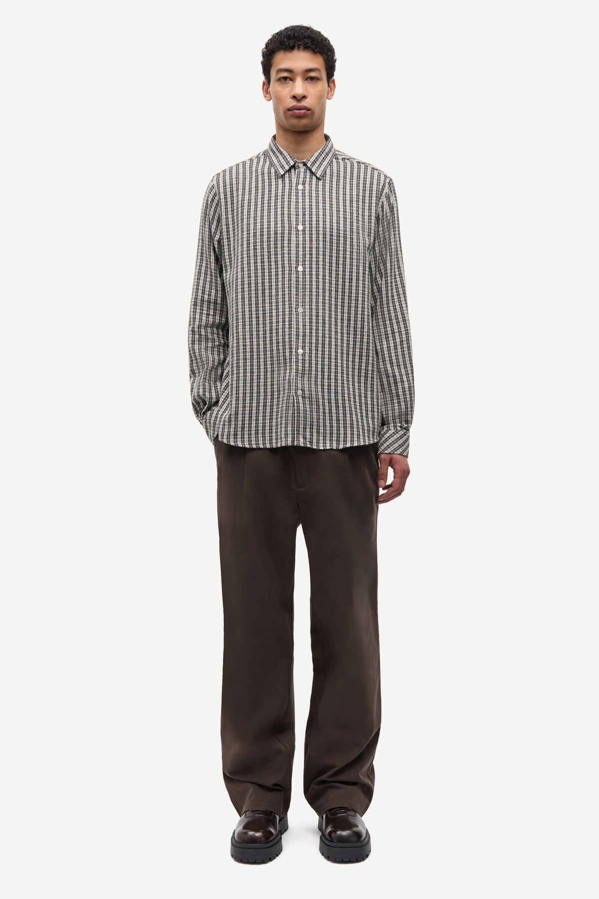 A man wearing a long-sleeve button-up shirt with vertical gray and white stripes, paired with dark brown trousers and dark brown shoes, standing against a light gray background.