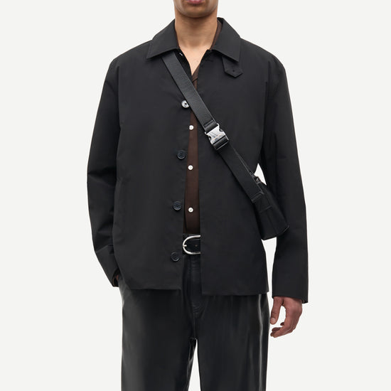 A man modeling a black button-front overshirt with a collar, worn over a brown shirt, paired with black trousers and a black crossbody bag with a silver buckle, against a light gray studio background.