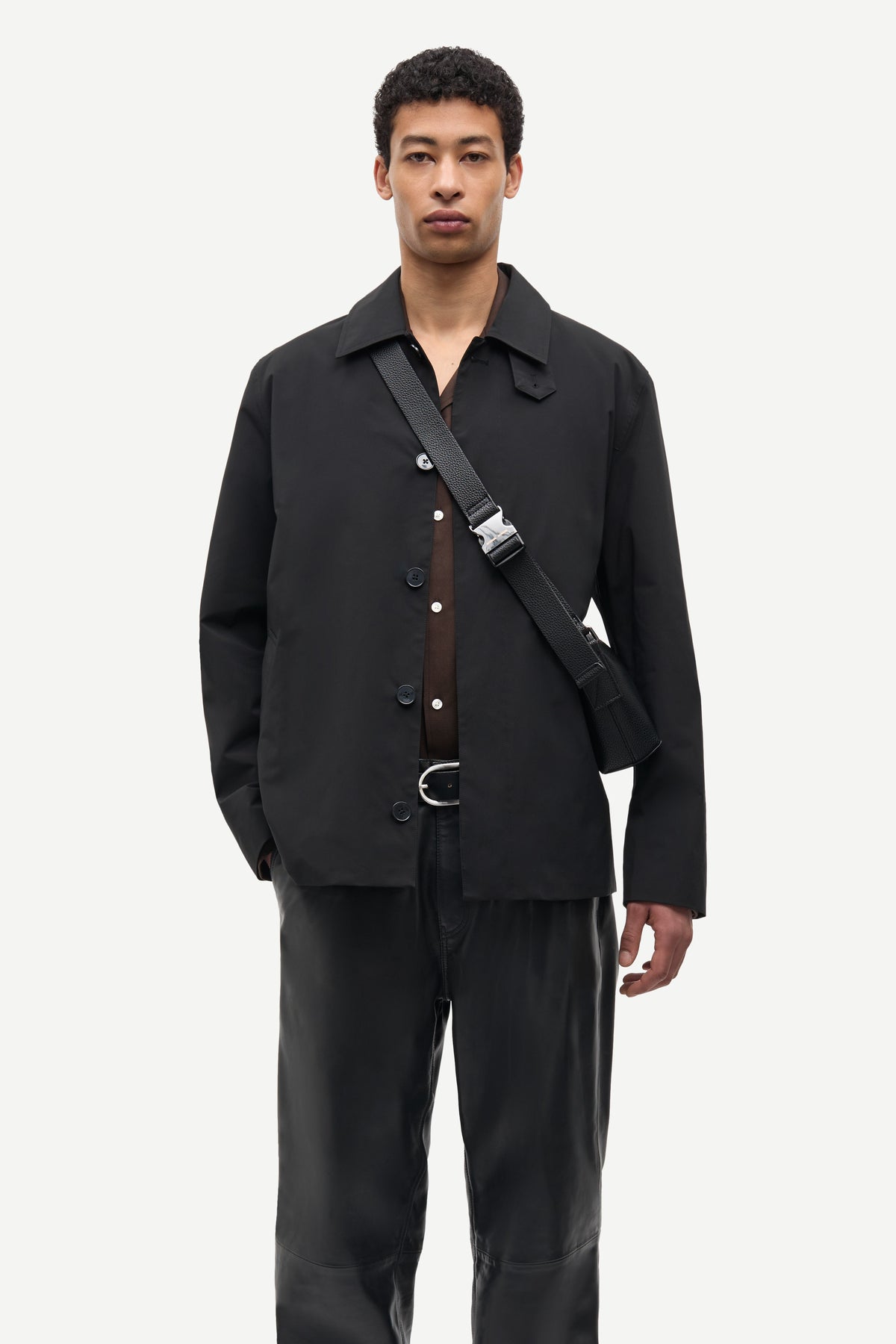 A man modeling a black button-front overshirt with a collar, worn over a brown shirt, paired with black trousers and a black crossbody bag with a silver buckle, against a light gray studio background.
