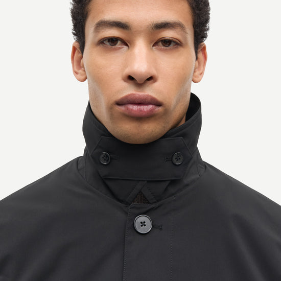 A close-up portrait of a man wearing a black high-neck jacket with a buttoned collar and a single visible button, set against a plain light gray background.