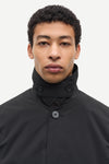 A close-up portrait of a man wearing a black high-neck jacket with a buttoned collar and a single visible button, set against a plain light gray background.