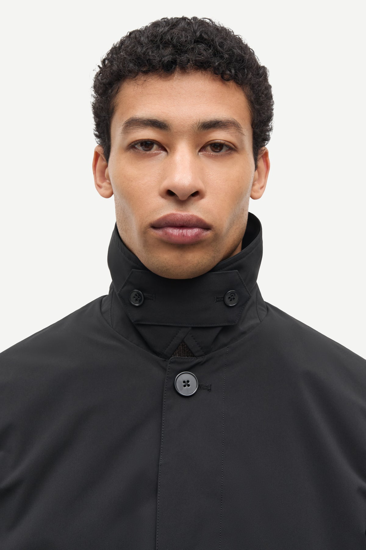 A close-up portrait of a man wearing a black high-neck jacket with a buttoned collar and a single visible button, set against a plain light gray background.