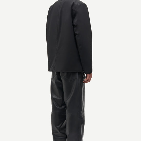 A man shown from the back wearing a black blazer and black trousers against a plain light gray studio background.