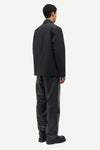 A man shown from the back wearing a black blazer and black trousers against a plain light gray studio background.