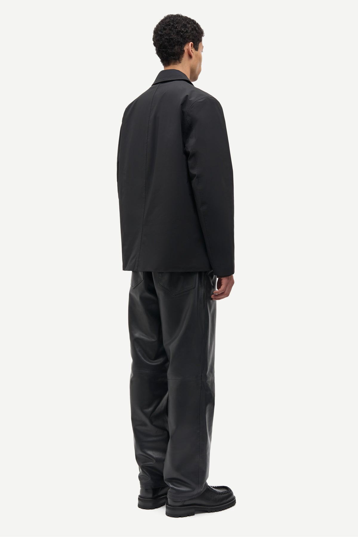 A man shown from the back wearing a black blazer and black trousers against a plain light gray studio background.
