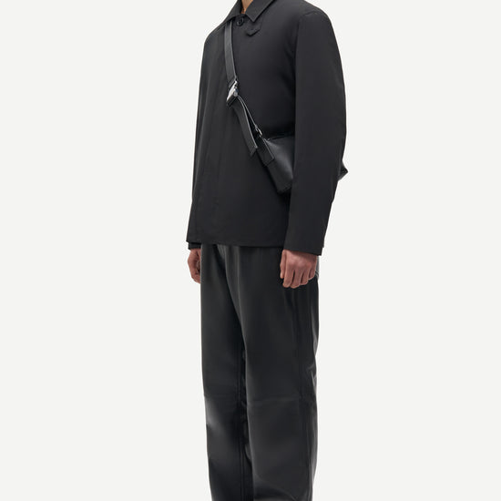 A man in a black long-sleeve collared shirt, black trousers, and black shoes stands in profile against a light gray studio background, wearing a black crossbody bag.