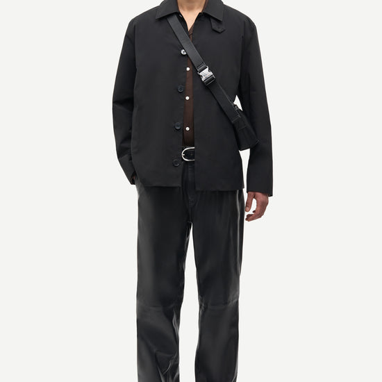 A male model stands against a light gray studio background wearing a black button-front overshirt, matching black trousers, black shoes, and a black crossbody bag with a silver buckle.
