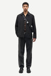 A male model stands against a light gray studio background wearing a black button-front overshirt, matching black trousers, black shoes, and a black crossbody bag with a silver buckle.