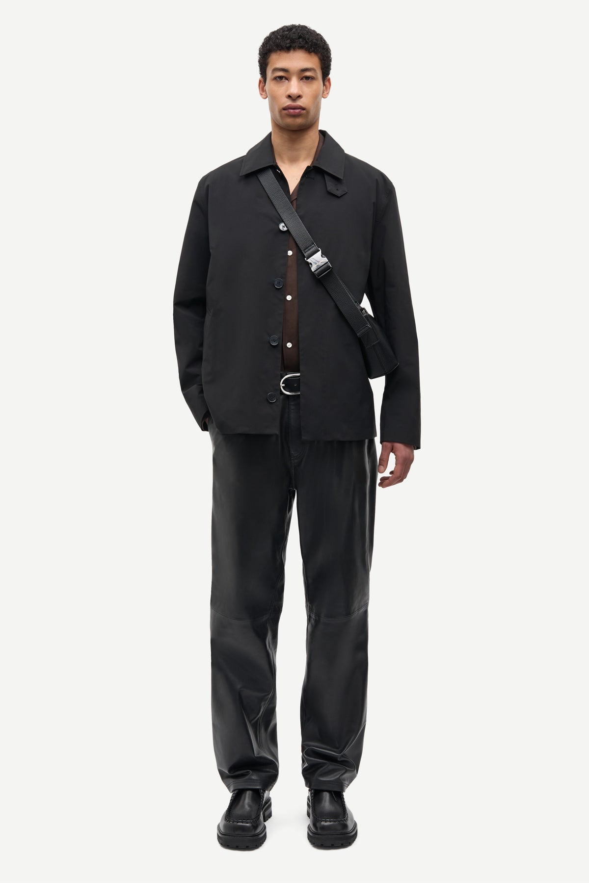 A male model stands against a light gray studio background wearing a black button-front overshirt, matching black trousers, black shoes, and a black crossbody bag with a silver buckle.