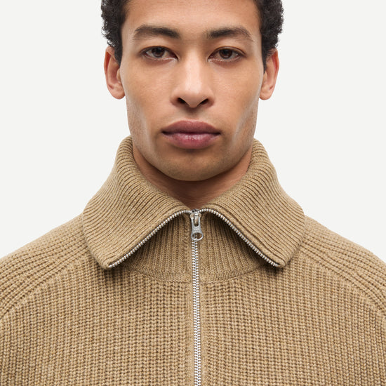 A close-up shot of a man wearing a beige ribbed knit quarter-zip sweater with a high collar, set against a plain light gray background.