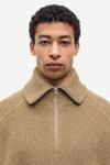 A close-up shot of a man wearing a beige ribbed knit quarter-zip sweater with a high collar, set against a plain light gray background.