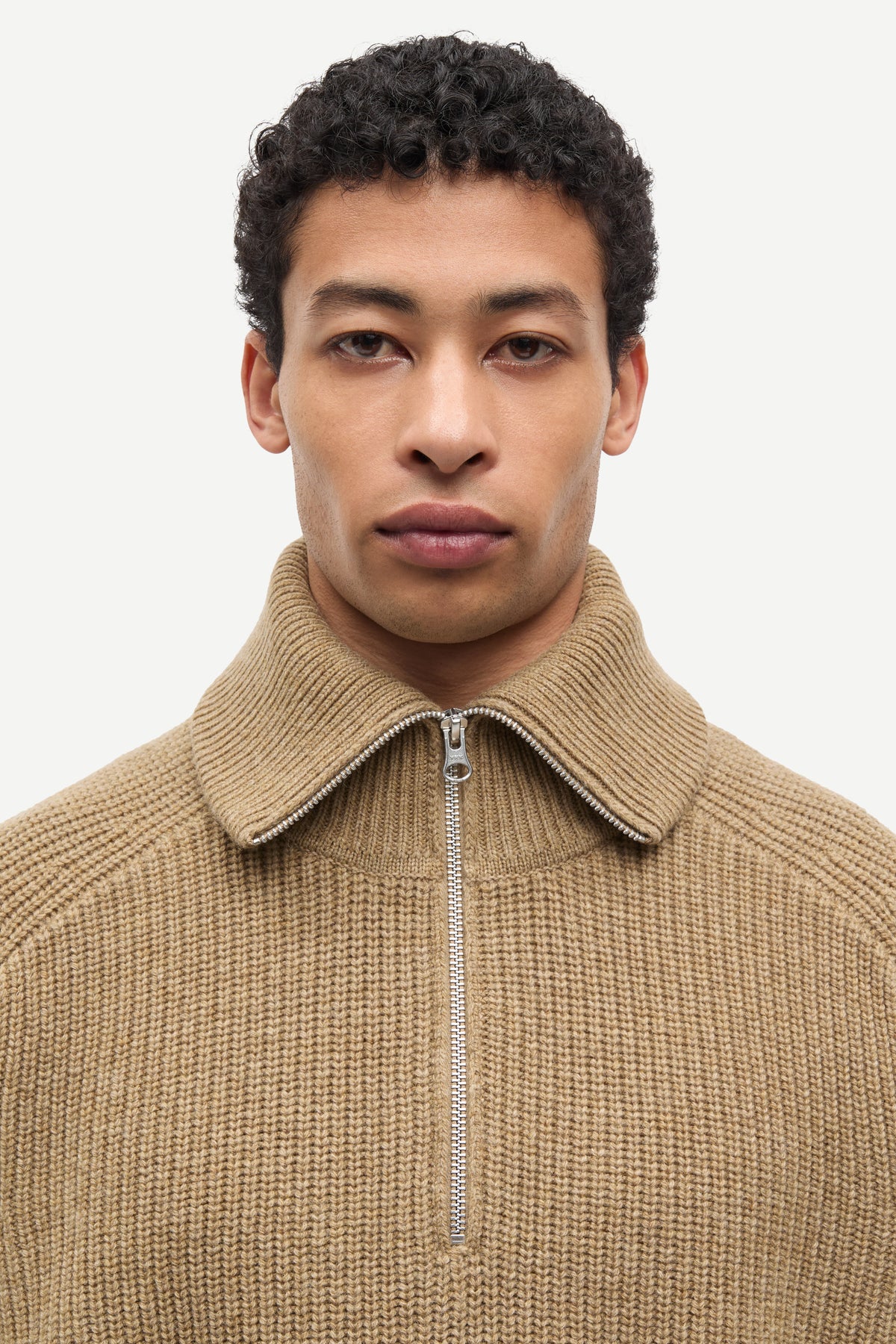 A close-up shot of a man wearing a beige ribbed knit quarter-zip sweater with a high collar, set against a plain light gray background.