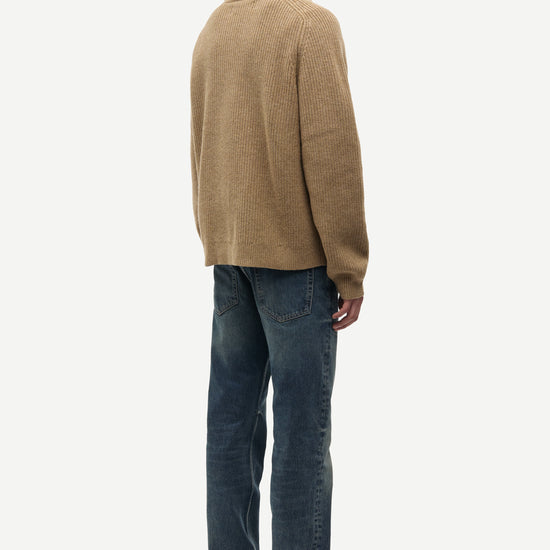 A man shown from the back wearing a tan knit sweater and dark blue jeans, standing against a plain light gray studio background.