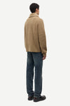 A man shown from the back wearing a tan knit sweater and dark blue jeans, standing against a plain light gray studio background.