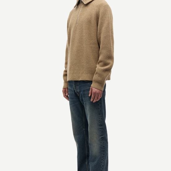 A man wearing a beige knit polo-neck sweater, dark blue jeans, and black leather shoes, standing in profile against a light gray studio background.