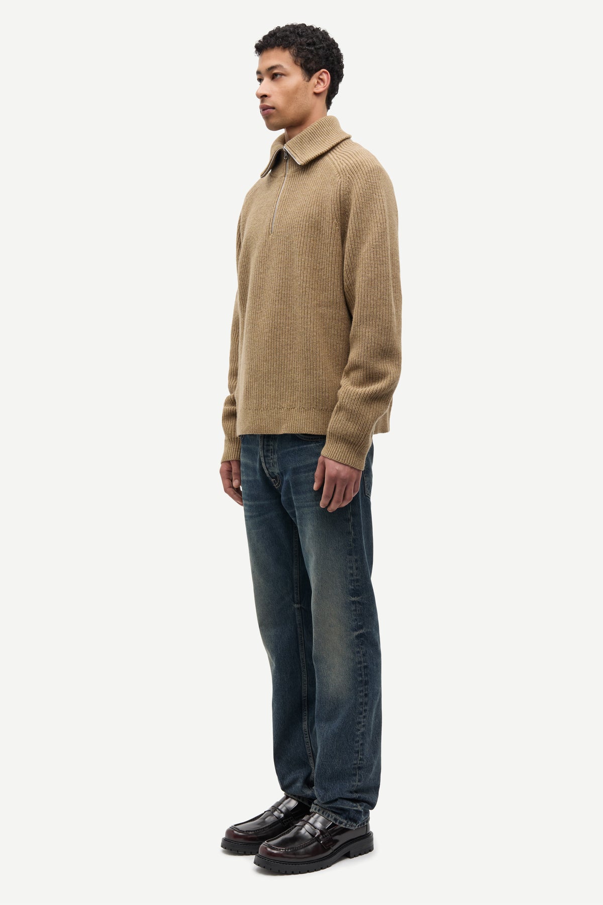 A man wearing a beige knit polo-neck sweater, dark blue jeans, and black leather shoes, standing in profile against a light gray studio background.