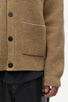 Close-up of a textured brown knit cardigan sweater with dark buttons and a front pocket, worn over a dark shirt and black pants.