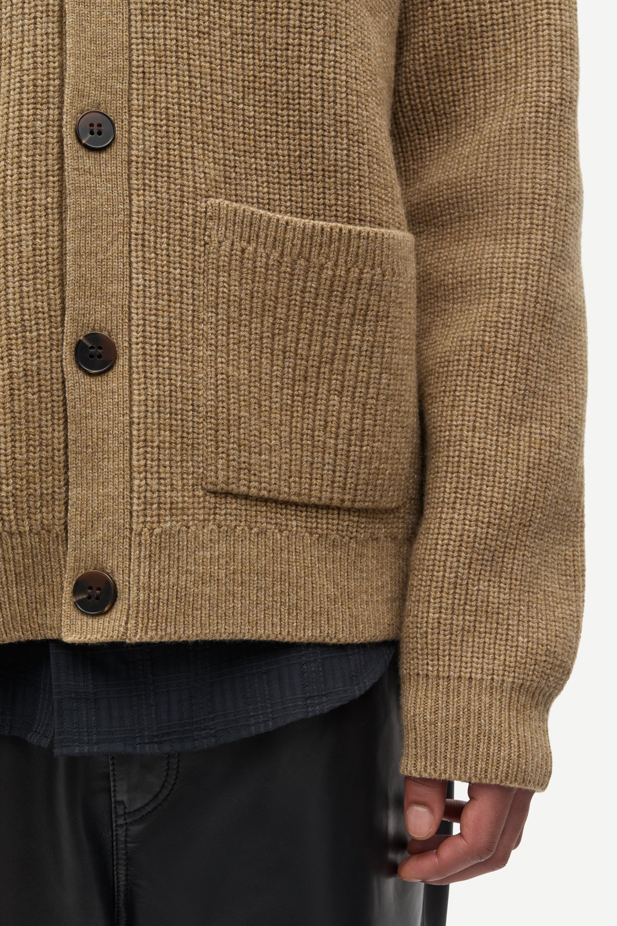 Close-up of a textured brown knit cardigan sweater with dark buttons and a front pocket, worn over a dark shirt and black pants.