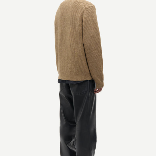 A man shown from the back wearing a tan knit sweater over a dark shirt and black wide-leg trousers, photographed against a plain light gray studio background.