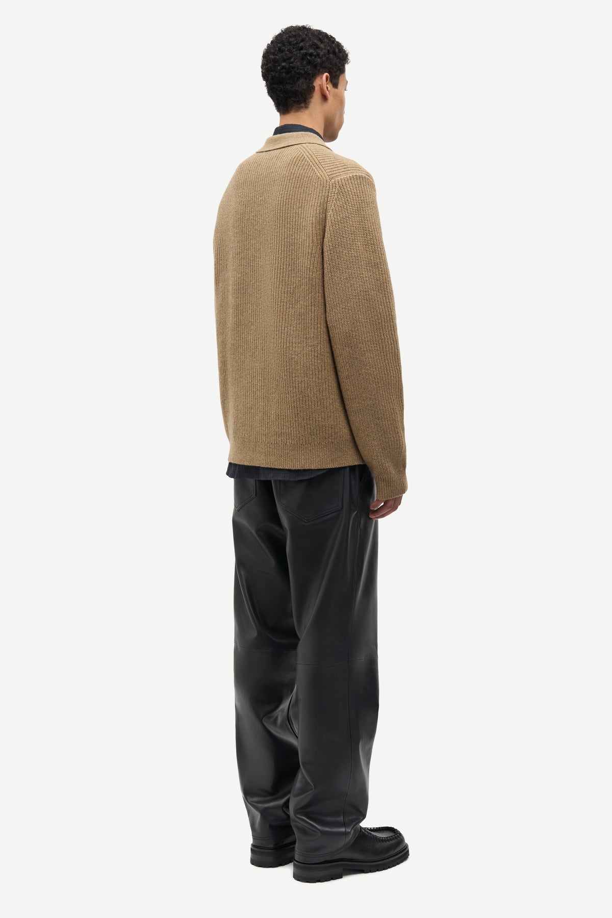 A man shown from the back wearing a tan knit sweater over a dark shirt and black wide-leg trousers, photographed against a plain light gray studio background.