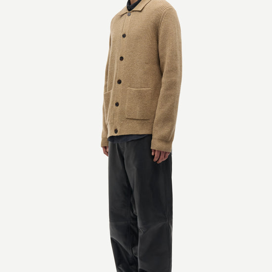 A man wearing a tan knit button-front cardigan with a pointed collar, paired with black trousers and black shoes, standing in profile against a plain light gray studio background.