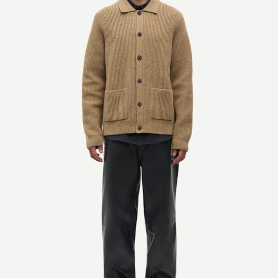 A male model stands against a light gray studio background wearing a tan ribbed knit button-front cardigan over a black collared shirt, paired with black trousers and black shoes.