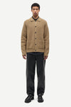 A male model stands against a light gray studio background wearing a tan ribbed knit button-front cardigan over a black collared shirt, paired with black trousers and black shoes.