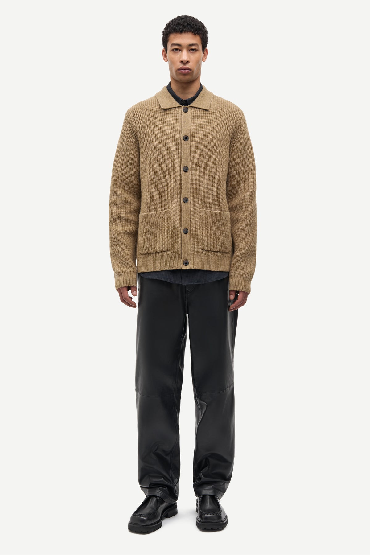 A male model stands against a light gray studio background wearing a tan ribbed knit button-front cardigan over a black collared shirt, paired with black trousers and black shoes.