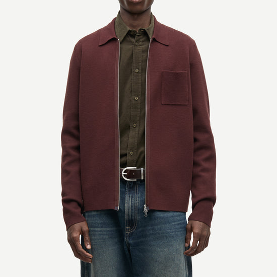 A male model wearing a burgundy zip-up jacket over an olive green button-down shirt and blue jeans, standing against a light gray studio background.