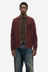 A male model wearing a burgundy zip-up jacket over an olive green button-down shirt and blue jeans, standing against a light gray studio background.