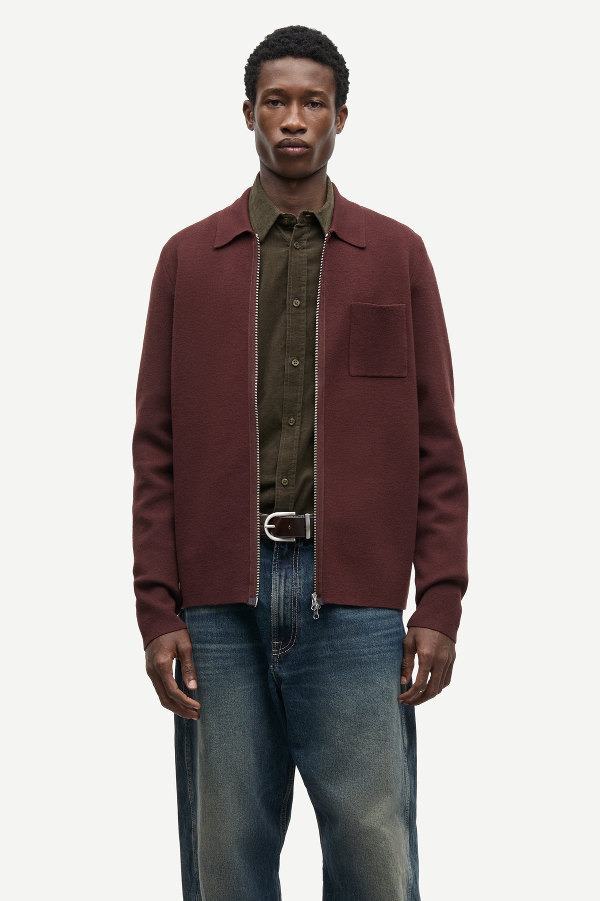A male model wearing a burgundy zip-up jacket over an olive green button-down shirt and blue jeans, standing against a light gray studio background.