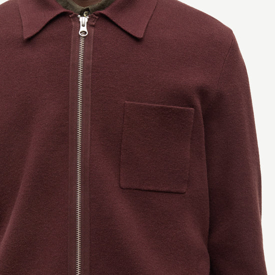 A close-up of a person wearing a burgundy zip-up collared jacket over a dark olive green shirt, against a plain light background.