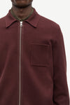 A close-up of a person wearing a burgundy zip-up collared jacket over a dark olive green shirt, against a plain light background.