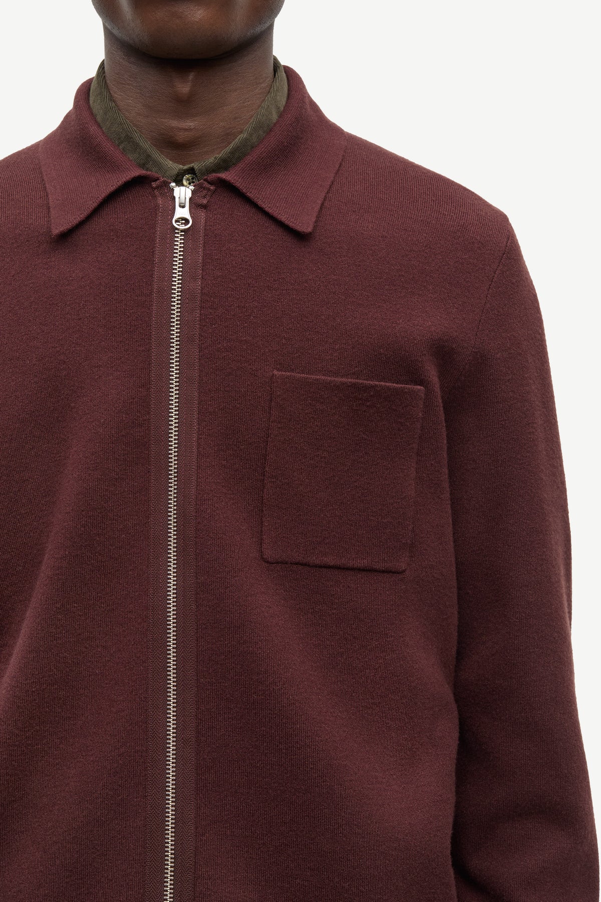 A close-up of a person wearing a burgundy zip-up collared jacket over a dark olive green shirt, against a plain light background.