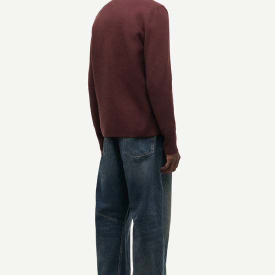 A man shown from the back wearing a dark burgundy knit sweater and wide-leg blue jeans, standing against a plain light gray studio background.