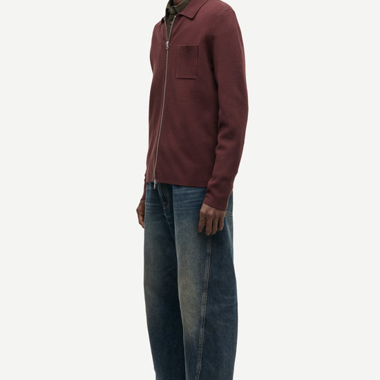 A man stands in profile wearing a burgundy zip-up jacket over a dark green collared shirt, blue jeans, and brown suede shoes, photographed against a clean off-white studio background.