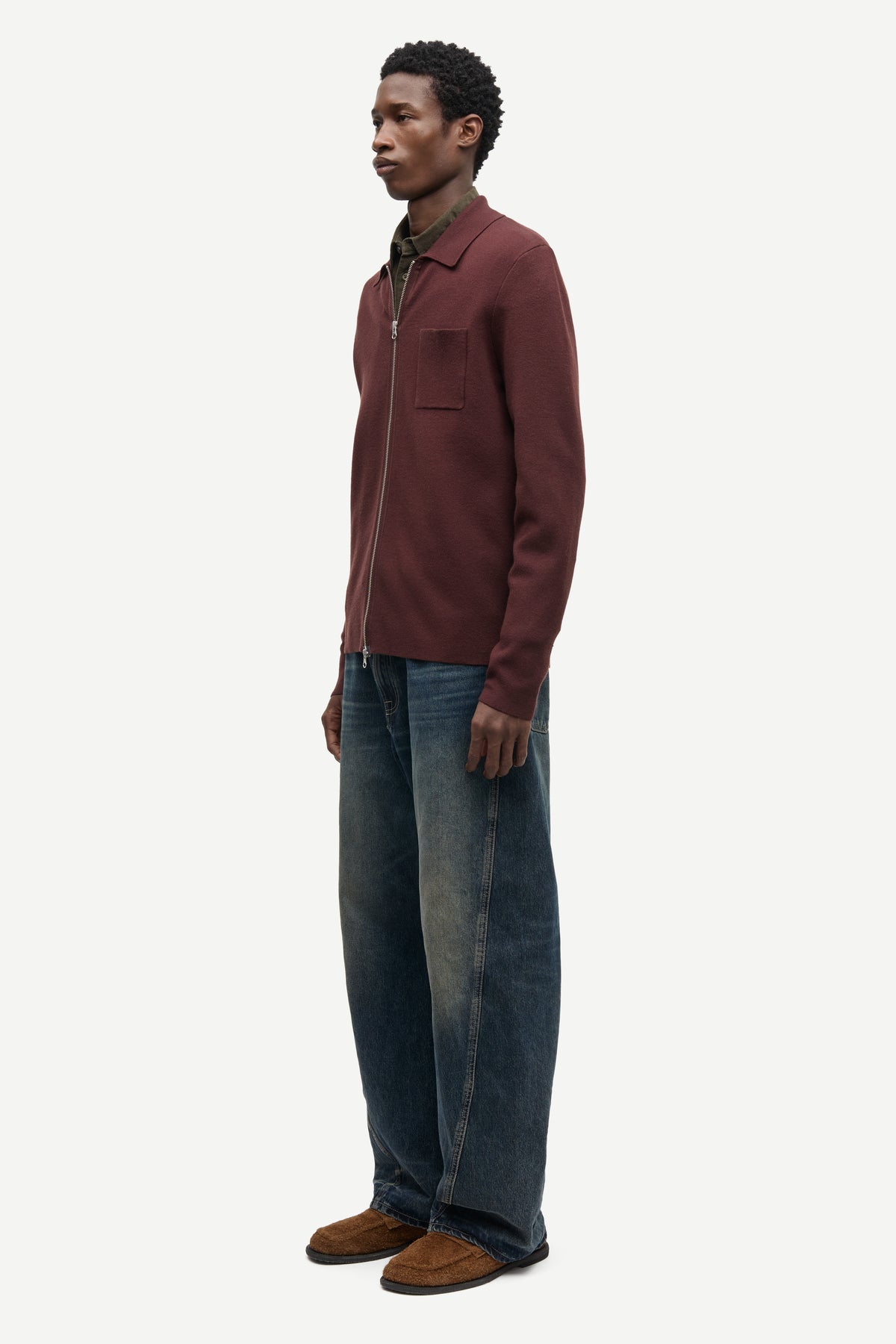 A man stands in profile wearing a burgundy zip-up jacket over a dark green collared shirt, blue jeans, and brown suede shoes, photographed against a clean off-white studio background.