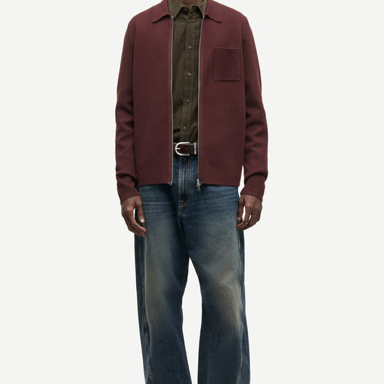 A male model stands against a light gray studio background wearing a burgundy overshirt over a dark olive green shirt, blue jeans, and brown shoes.