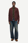 A male model stands against a light gray studio background wearing a burgundy overshirt over a dark olive green shirt, blue jeans, and brown shoes.
