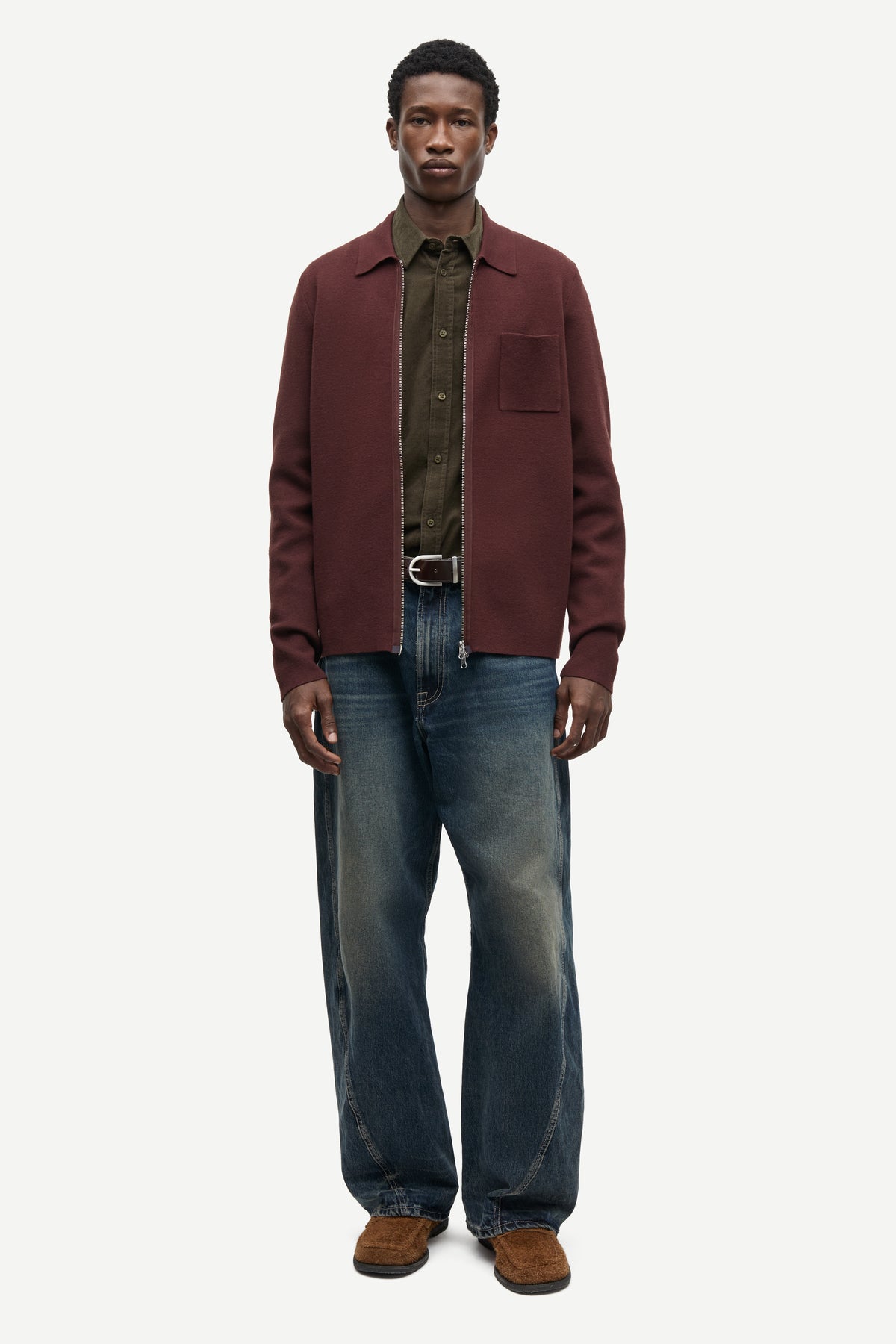 A male model stands against a light gray studio background wearing a burgundy overshirt over a dark olive green shirt, blue jeans, and brown shoes.