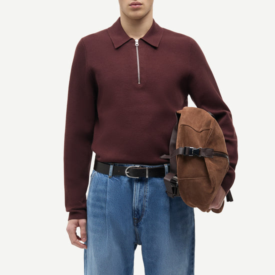 A male model wearing a burgundy long-sleeve quarter-zip polo sweater, blue jeans, and a black belt, holding a brown suede crossbody bag against a plain light gray background.