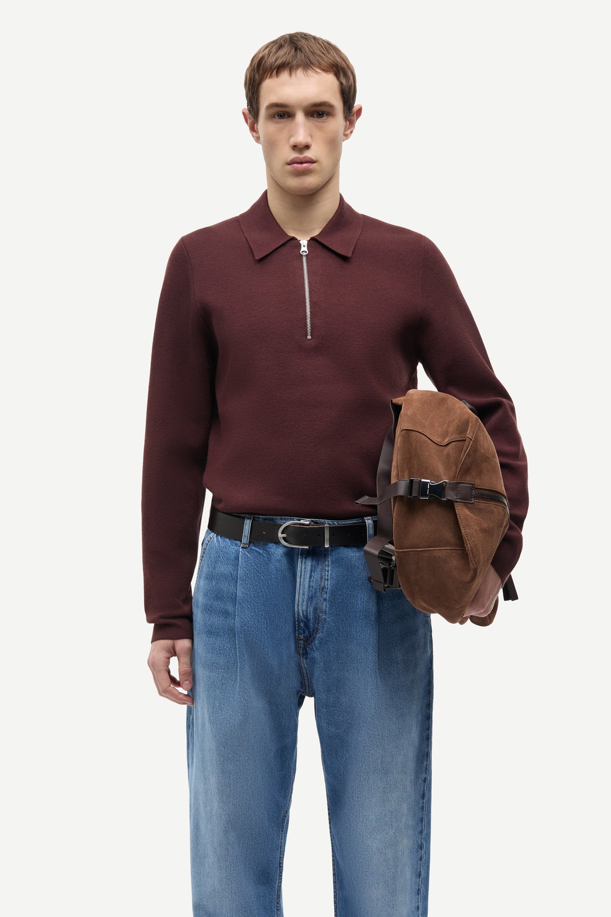 A male model wearing a burgundy long-sleeve quarter-zip polo sweater, blue jeans, and a black belt, holding a brown suede crossbody bag against a plain light gray background.