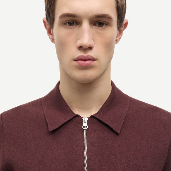 A man wearing a burgundy half-zip knit polo sweater, shown from the chest up against a plain light gray background.