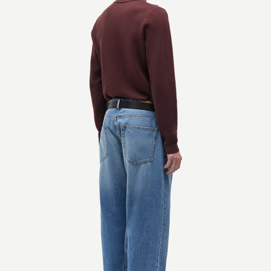 A man shown from the back wearing a dark burgundy long-sleeve knit polo sweater, light blue wide-leg jeans, a black belt, and brown suede shoes, against a plain light gray studio background.