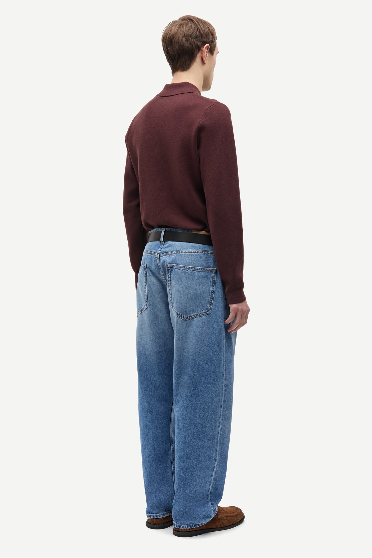 A man shown from the back wearing a dark burgundy long-sleeve knit polo sweater, light blue wide-leg jeans, a black belt, and brown suede shoes, against a plain light gray studio background.