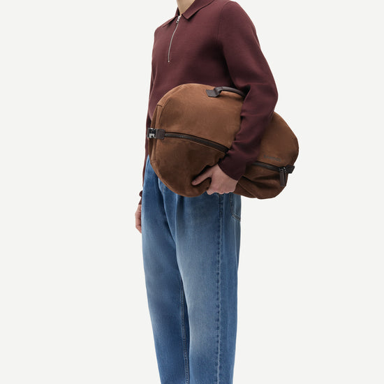 A man in a maroon half-zip pullover, blue jeans, and brown suede shoes is shown in profile holding a brown suede shoulder bag with black trim against a light gray studio background.